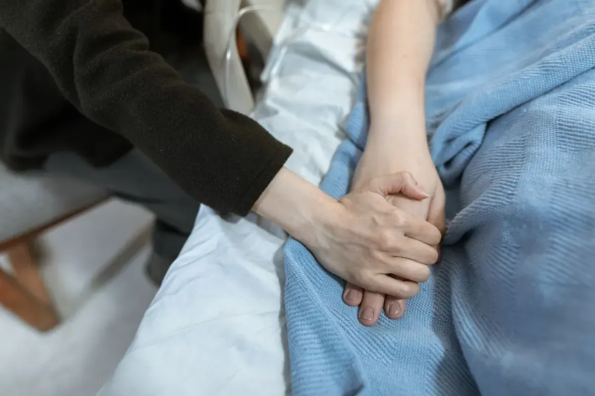 A comforting hand holding a patient’s hand in a hospital bed during illness.